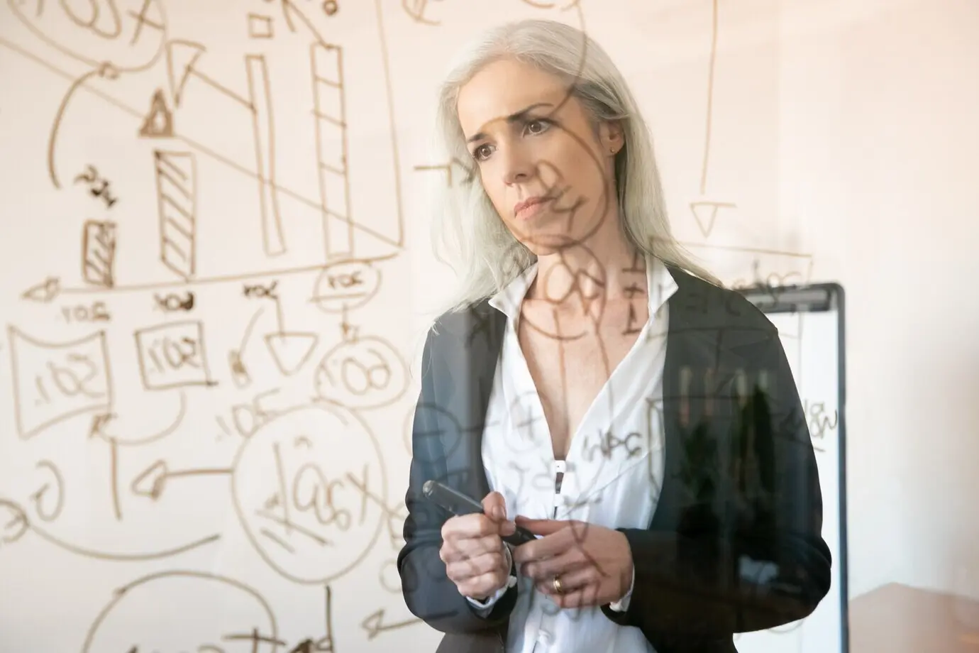 Седовласая бизнес-леди изучает статистические данные и размышляет. Серьезная, опытная, вдумчивая женщина-менеджер держит маркер и стоит в офисном помещении. Тематика: стратегия, бизнес и управление.